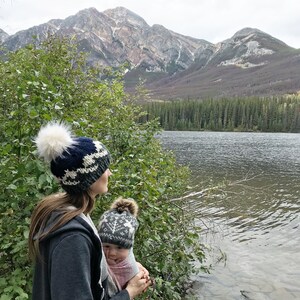 Puede incluir: Una mujer con un gorro de punto azul y blanco con un pomp&oacute;n blanco sostiene a un beb&eacute; que lleva un gorro de punto gris y blanco con un pomp&oacute;n blanco. Est&aacute;n de pie junto a un lago con monta&ntilde;as al fondo.