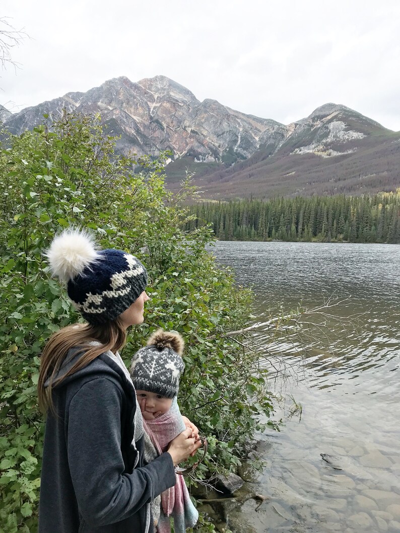 Puede incluir: Una mujer con un gorro de punto azul y blanco con un pomp&oacute;n blanco sostiene a un beb&eacute; que lleva un gorro de punto gris y blanco con un pomp&oacute;n blanco. Est&aacute;n de pie junto a un lago con monta&ntilde;as al fondo.