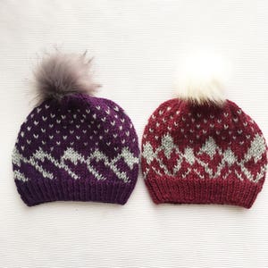 May include: Two knitted winter hats with pom poms. One hat is purple with a gray mountain pattern and the other is burgundy with a gray mountain pattern.