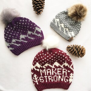 May include: Three knitted winter hats with pom poms. The hats are purple, gray, and burgundy with white accents. The burgundy hat has the words "Maker Strong" knitted into the design.