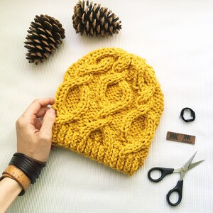 May include: A yellow knitted beanie with a cable knit pattern. The beanie is on a white surface with pine cones and a pair of scissors.
