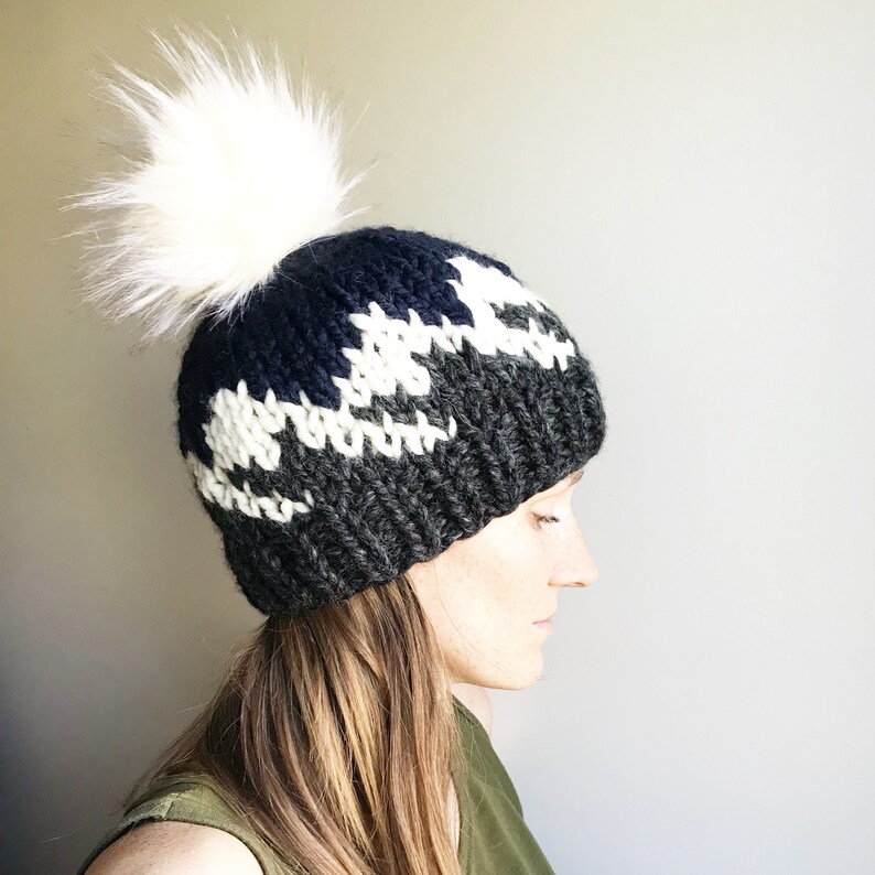 Puede incluir: Un gorro de punto negro, blanco y gris con un pomp&oacute;n de piel sint&eacute;tica blanca. El gorro tiene un patr&oacute;n geom&eacute;trico.