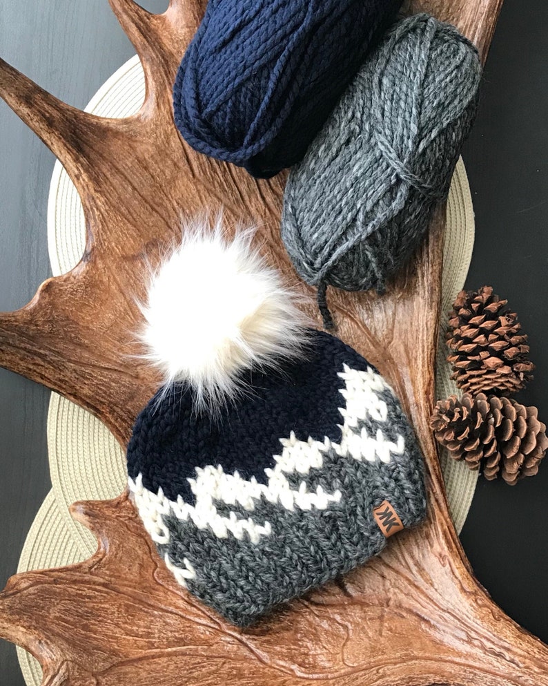 Puede incluir: Un gorro de punto con un pomp&oacute;n blanco, que presenta un patr&oacute;n de monta&ntilde;a azul y gris. El gorro tiene una etiqueta de cuero con las iniciales "MK".