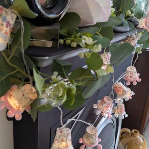 May include: A fireplace mantel decorated with a string of floral lights. The lights are cup-shaped with pink pom-poms. Eucalyptus and white flowers are arranged around the lights. A large candle sits on the mantelpiece.