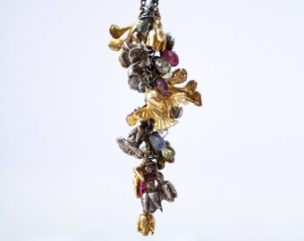 May include: A long, delicate necklace with a silver and gold pendant. The pendant features a cluster of small, intricately crafted leaves and flowers, adorned with colourful gemstones.