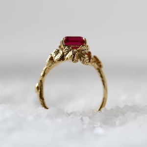 May include: A gold ring with a red gemstone and leaf-like details. The ring is set on a white background.