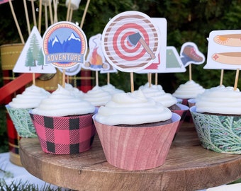 May include: A wooden cake stand with cupcakes decorated with camping-themed toppers. The toppers include a campfire, a tent, a pair of hiking boots, a canoe, an axe, and a mountain range with the word "ADVENTURE".