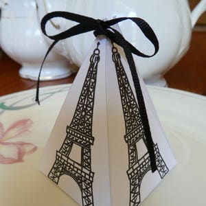 Puede incluir: Una caja de papel blanco en forma de pirámide con una cinta negra atada alrededor de la parte superior. La caja está decorada con una ilustración en blanco y negro de la Torre Eiffel.