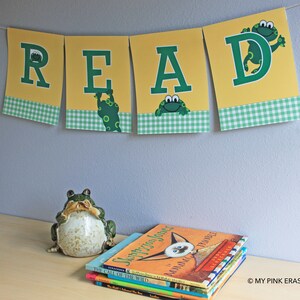 May include: A yellow and green banner with the word "READ" in large green letters. Each letter has a small green frog peeking out from behind it. The banner is decorated with a green and white checkered pattern.