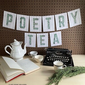 May include: A white teapot, a book, a typewriter, and four teacups are arranged on a light wood surface. A banner with the words "Poetry Tea" hangs above the scene. The banner is made of white paper with a green leafy pattern.