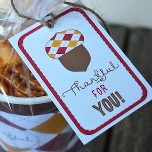 Può includere: Un'etichetta regalo bianca con un bordo rosso e un'illustrazione di una ghianda marrone. L'etichetta dice "Grazie per te!" ed è legata con uno spago a un sacchetto di plastica trasparente pieno di uno spuntino.