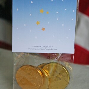 Puede incluir: Una bolsa de pl&aacute;stico transparente que contiene varias monedas de chocolate doradas. La bolsa est&aacute; sellada con una tarjeta que presenta un dise&ntilde;o de cielo azul degradado con puntos blancos y estrellas amarillas. El texto en la tarjeta dice "MY PINK ERASER 2017" y una direcci&oacute;n web.