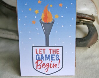 Puede incluir: Una tarjeta azul y blanca con un escudo rojo, blanco y azul que dice "Let the Games Begin!"  Una antorcha con una llama está por encima del escudo.  Hay estrellas amarillas y puntos blancos en el fondo azul.  La tarjeta está sobre una superficie blanca.