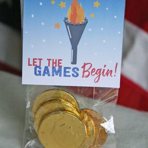 Puede incluir: Una bolsa transparente llena de monedas de chocolate doradas. La bolsa est&aacute; coronada con una tarjeta con un gr&aacute;fico de una antorcha y el texto "LET THE GAMES Begin!" sobre un fondo de cielo azul con estrellas.