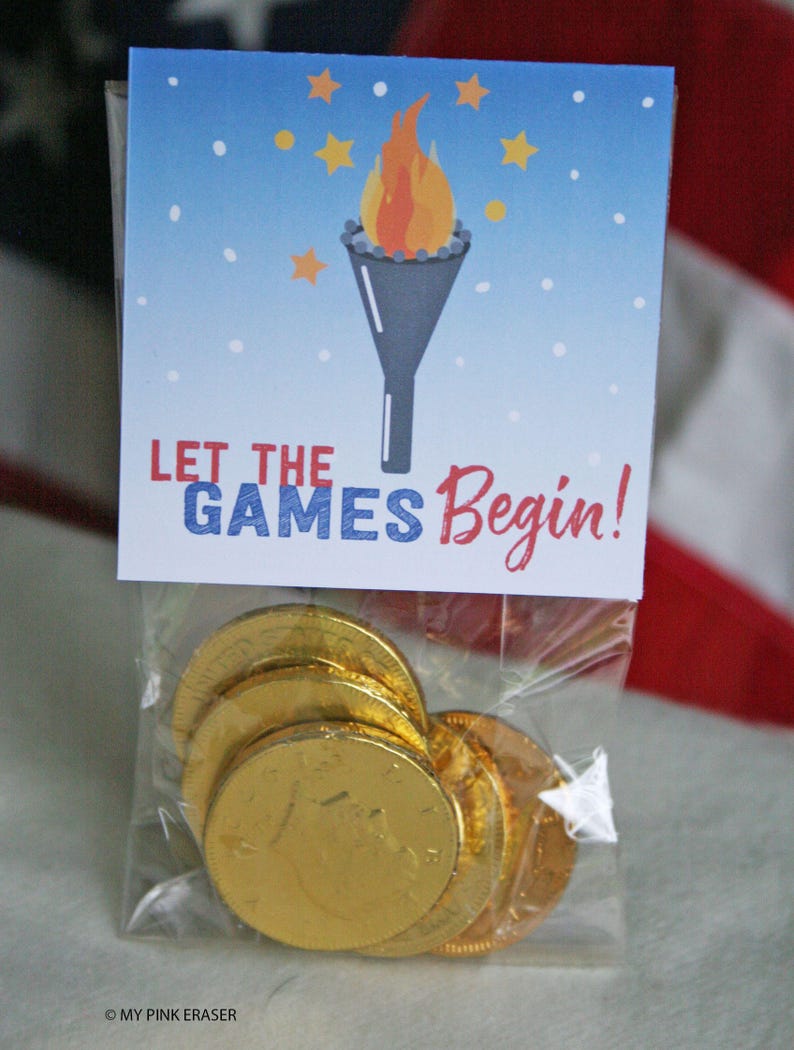 Puede incluir: Una bolsa transparente llena de monedas de chocolate doradas. La bolsa est&aacute; coronada con una tarjeta con un gr&aacute;fico de una antorcha y el texto "LET THE GAMES Begin!" sobre un fondo de cielo azul con estrellas.