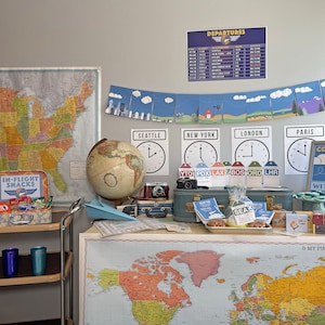 May include: A travel-themed party setup with a blue suitcase, a globe, maps of the United States and the world, and signs that say "Gate Check-In", "In-Flight Snacks", "Come Fly With Me", and "Baggage Claim".