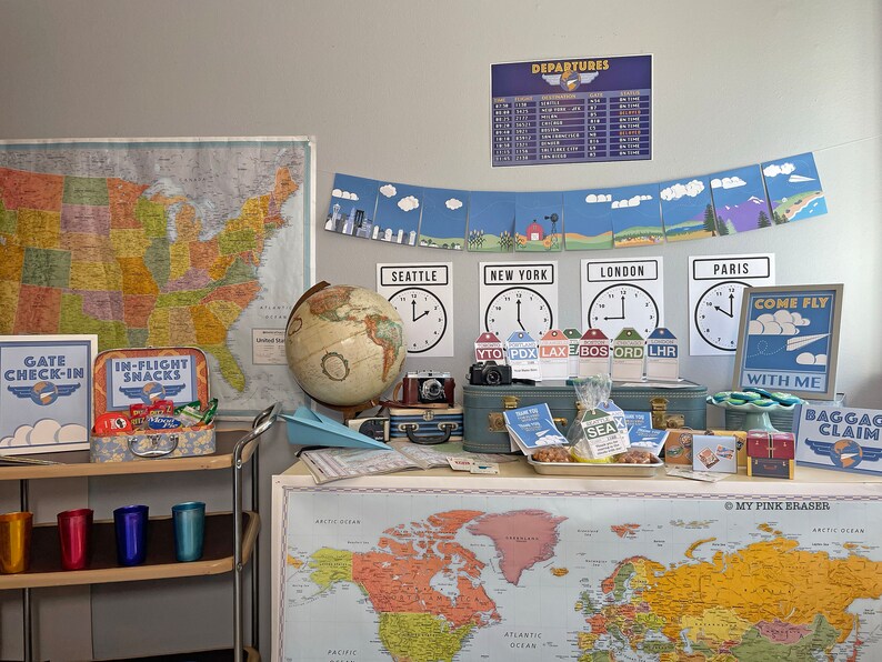 May include: A travel-themed party setup with a blue suitcase, a globe, maps of the United States and the world, and signs that say "Gate Check-In", "In-Flight Snacks", "Come Fly With Me", and "Baggage Claim".