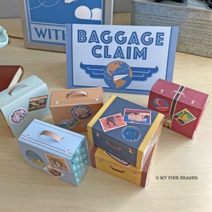 May include: A set of six miniature luggage boxes with various travel-themed designs. The boxes are blue, yellow, and red with white accents. The boxes are arranged on a wooden surface with a sign that reads "Baggage Claim".
