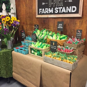 May include: A farm stand themed party table with a brown paper tablecloth. The table is decorated with various treats, including popcorn in green bags, red candy in baskets, and caramel corn in wooden crates. The table is decorated with signs that say "You're Really Amazing!" and "We Carrot About You!"