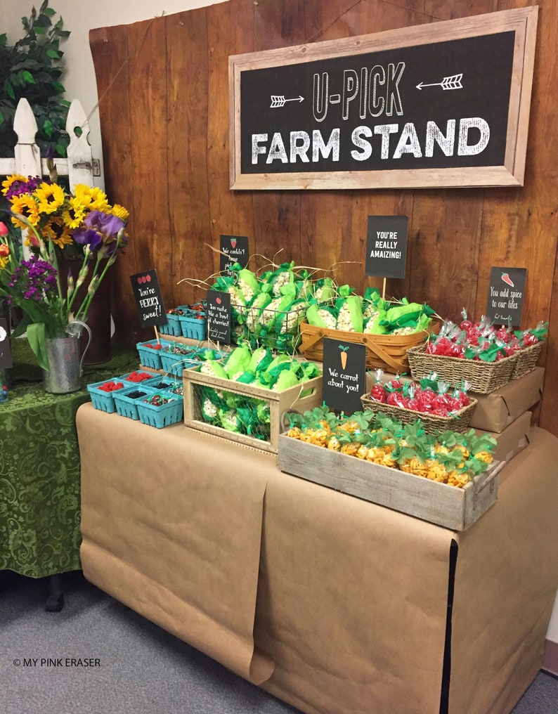 May include: A farm stand themed party table with a brown paper tablecloth. The table is decorated with various treats, including popcorn in green bags, red candy in baskets, and caramel corn in wooden crates. The table is decorated with signs that say "You're Really Amazing!" and "We Carrot About You!"