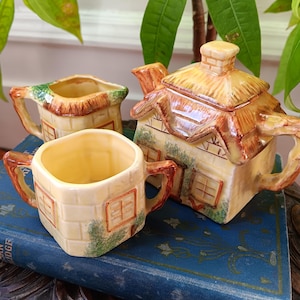 May include: A vintage ceramic tea set comprising a teapot, creamer jug, and sugar bowl, each shaped like a quaint cottage. The teapot has a thatched roof and brick-effect walls. The set is in beige, brown, and green, creating a charming, rustic feel.