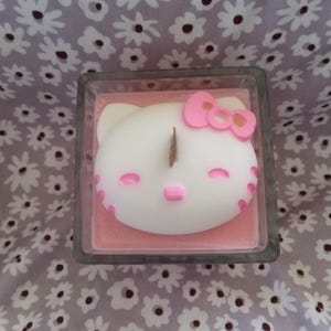 Puede incluir: Una vela en recipiente de vidrio cuadrado relleno de cera rosa, con una figura de cera blanca en forma de gato que tiene un lazo rosa, ojos rosas y nariz rosa. Una mecha de madera sobresale del centro de la figura de cera blanca.