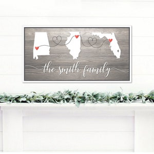 May include: A gray wood sign with white outlines of the states of Alabama, Illinois, and Florida. Red hearts are placed on each state. The text "the smith family" is written in white cursive script below the states.