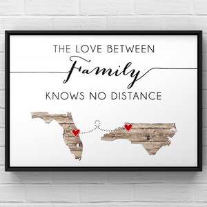 May include: Framed print with the text "THE LOVE BETWEEN FAMILY KNOWS NO DISTANCE." Features two state outlines with red heart markers connected by a dotted line. The state outlines have a wood grain texture.