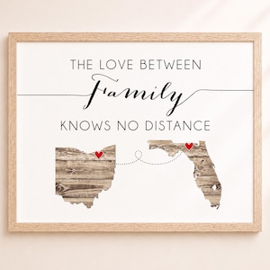 May include: A framed wall art print featuring the text THE LOVE BETWEEN Family KNOWS NO DISTANCE. Below the text are silhouettes of Ohio and Florida with a wood grain texture, connected by a dotted line with a red heart on each state.