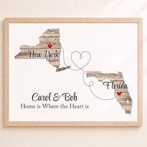 May include: A framed print featuring maps of New York and Florida with a wood grain texture. A black line connects the two states, forming a heart shape in the center. Red hearts mark locations, with the text Carol & Bob and Home is Where the Heart is.