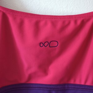 Puede incluir: Primer plano de un top deportivo magenta y morado. El top tiene un escote redondo y un peque&ntilde;o logotipo azul oscuro. El top est&aacute; hecho de una tela lisa, probablemente dise&ntilde;ada para absorber la humedad, y es probable que sea para deportes o fitness.