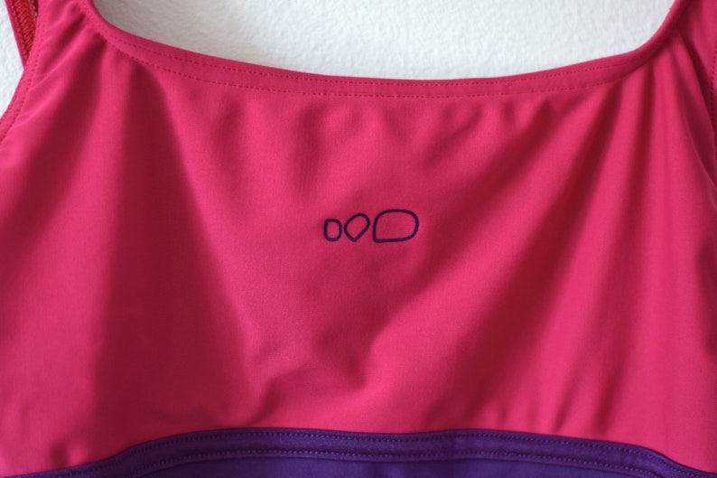 Puede incluir: Primer plano de un top deportivo magenta y morado. El top tiene un escote redondo y un peque&ntilde;o logotipo azul oscuro. El top est&aacute; hecho de una tela lisa, probablemente dise&ntilde;ada para absorber la humedad, y es probable que sea para deportes o fitness.