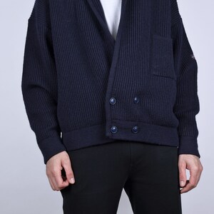 May include: A dark blue knitted cardigan with a shawl collar, two button closures, and a patch pocket on the left side. The cardigan is made of a ribbed fabric.