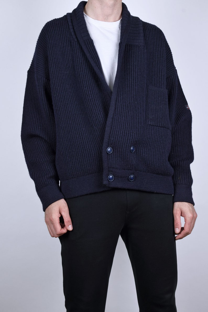 May include: A dark blue knitted cardigan with a shawl collar, two button closures, and a patch pocket on the left side. The cardigan is made of a ribbed fabric.