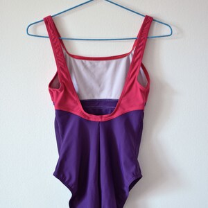 Puede incluir: Un ba&ntilde;ador de estilo vintage con un dise&ntilde;o de bloques de color. La parte superior es rosa y roja, mientras que la inferior es de color p&uacute;rpura oscuro. Cuelga de una percha azul contra una pared blanca. Un ba&ntilde;ador retro.