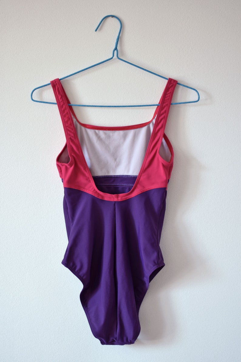 Puede incluir: Un ba&ntilde;ador de estilo vintage con un dise&ntilde;o de bloques de color. La parte superior es rosa y roja, mientras que la inferior es de color p&uacute;rpura oscuro. Cuelga de una percha azul contra una pared blanca. Un ba&ntilde;ador retro.