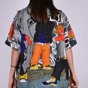 K&ouml;nnte beinhalten: Ein kurz&auml;rmeliges Hemd mit Kn&ouml;pfen und einem bunten Cartoon-Druck von drei Personen in Streetwear. Das Hemd ist grau mit schwarzen, orangen, blauen, roten und gelben Akzenten.