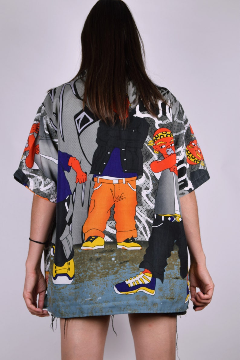 K&ouml;nnte beinhalten: Ein kurz&auml;rmeliges Hemd mit Kn&ouml;pfen und einem bunten Cartoon-Druck von drei Personen in Streetwear. Das Hemd ist grau mit schwarzen, orangen, blauen, roten und gelben Akzenten.