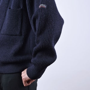May include: A dark blue knitted cardigan with a ribbed texture and a small patch on the sleeve that reads "RIPPER".