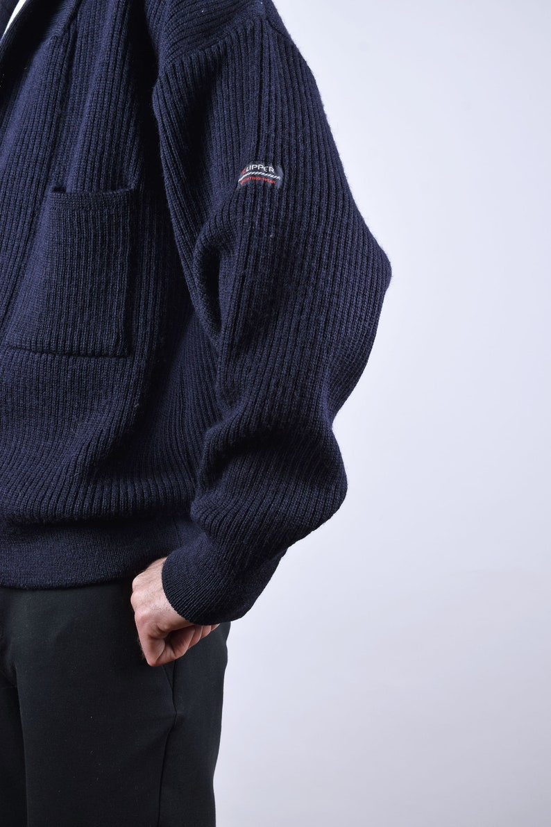 May include: A dark blue knitted cardigan with a ribbed texture and a small patch on the sleeve that reads "RIPPER".