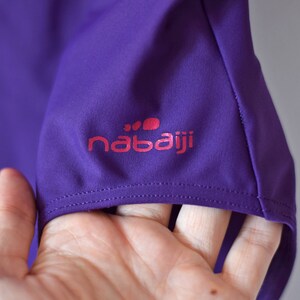 Puede incluir: Primer plano de una manga de camiseta deportiva morada. La manga presenta el logotipo rosa "nabaiji". La tela es de un morado liso y vibrante. La costura es visible a lo largo del dobladillo de la manga.