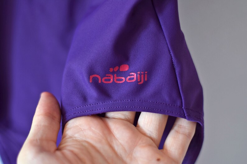 Puede incluir: Primer plano de una manga de camiseta deportiva morada. La manga presenta el logotipo rosa "nabaiji". La tela es de un morado liso y vibrante. La costura es visible a lo largo del dobladillo de la manga.