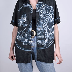 May include: A black button-up shirt with a short sleeve featuring a white tiger and a dragon in a yin-yang design. The shirt has a black background with blue and white details.