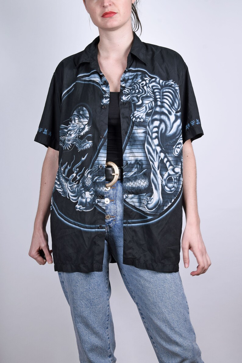 May include: A black button-up shirt with a short sleeve featuring a white tiger and a dragon in a yin-yang design. The shirt has a black background with blue and white details.