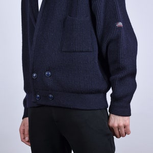 May include: A dark blue knitted cardigan with a shawl collar, two button closures, and a patch pocket on the left side. The cardigan has a ribbed texture and a brand label on the right sleeve.