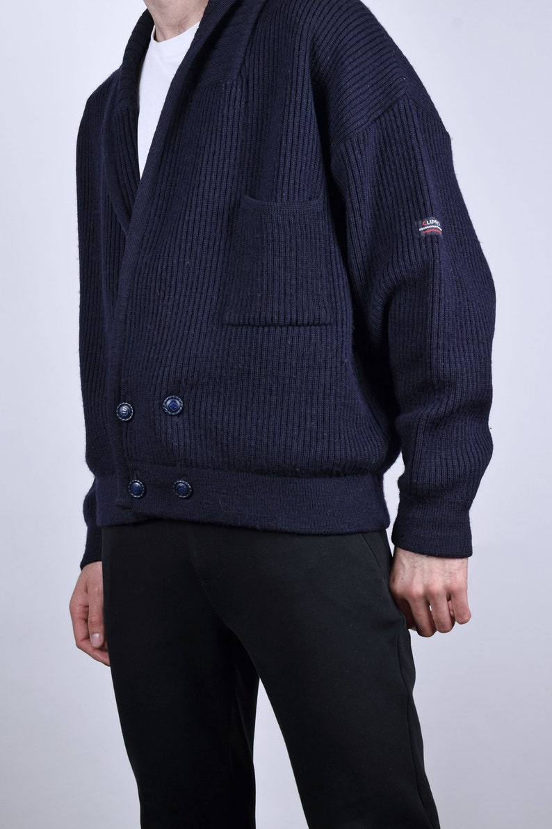 May include: A dark blue knitted cardigan with a shawl collar, two button closures, and a patch pocket on the left side. The cardigan has a ribbed texture and a brand label on the right sleeve.