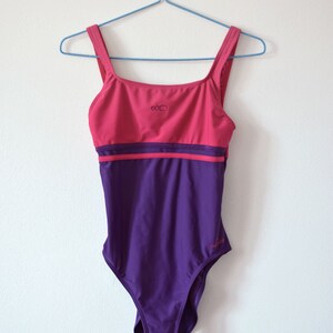 Puede incluir: Un ba&ntilde;ador de dos tonos en rosa y morado, colgado de una percha azul. La parte superior es rosa con un peque&ntilde;o logotipo y la parte inferior es morada. El ba&ntilde;ador tiene un escote cuadrado y tirantes anchos.