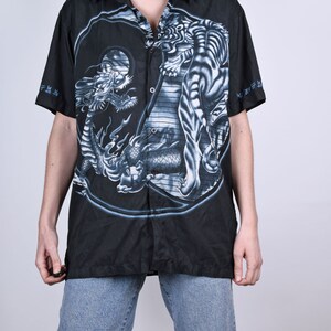 May include: Black button-up shirt with a white tiger and dragon print. The print features a yin-yang symbol with the tiger on one side and the dragon on the other. The shirt has short sleeves and a collared neckline.