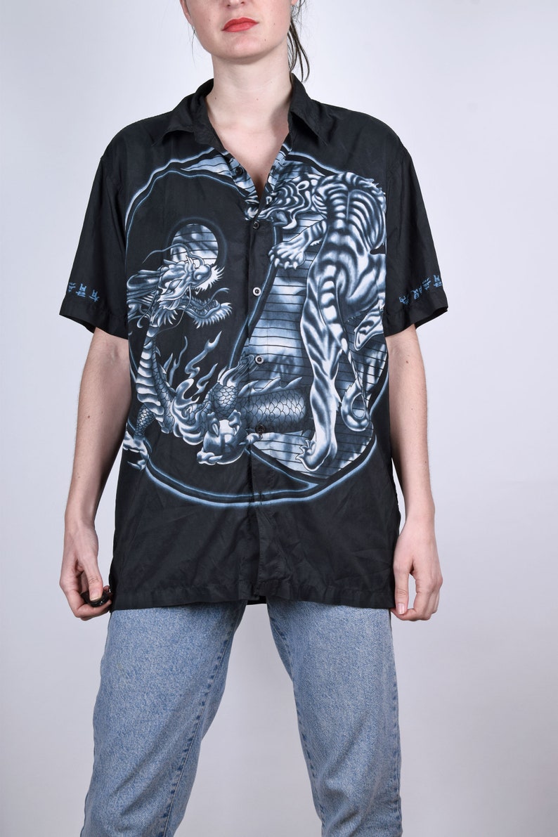 May include: Black button-up shirt with a white tiger and dragon print. The print features a yin-yang symbol with the tiger on one side and the dragon on the other. The shirt has short sleeves and a collared neckline.