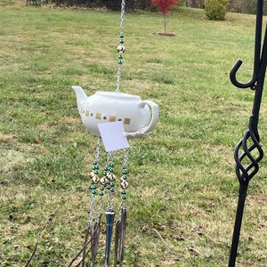 Puede incluir: Un carillón de té blanco con detalles dorados cuelga de un soporte de metal negro en un campo de hierba. El carillón está hecho de cucharas y cuentas.